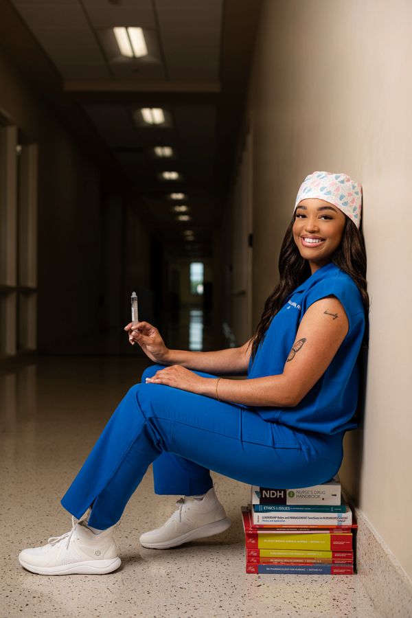 Smiling nurse in blue scrubs holding a syringe, sitting on nursing textbooks.