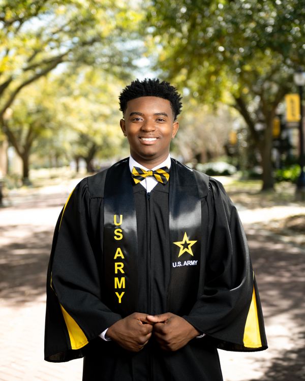 Young man in US Army graduation gown smiling outdoors.