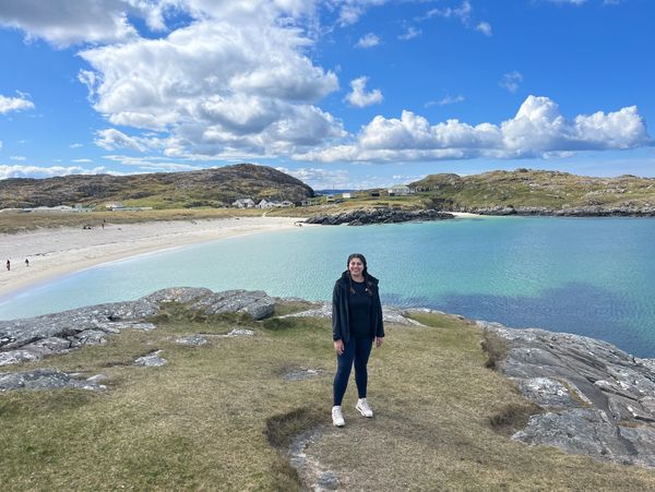 A woman stands on a grassy rock by a turquoise bay under a blue sky with clouds.