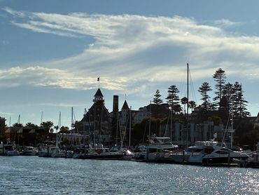Sailboats docked at a marina with a historic building and trees in the background under a partly cloudy sky.