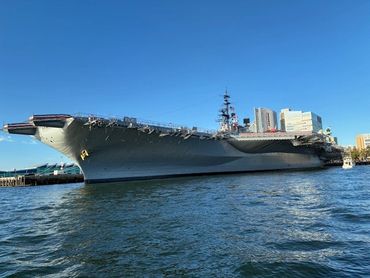Large aircraft carrier docked near city buildings under clear blue sky.