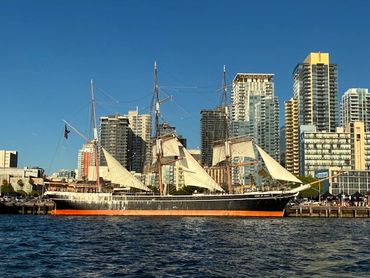Historic sailing ship docked by modern city skyscrapers under clear blue sky.