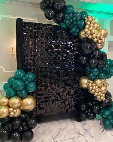 Black sequin backdrop with green, gold, and black balloon arch in an elegant room.