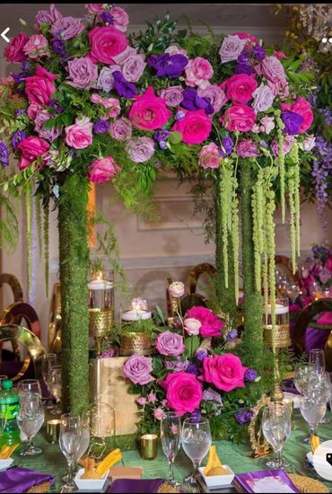 Lavish floral centerpiece with vibrant pink and purple roses, adorned with hanging greenery and candles.