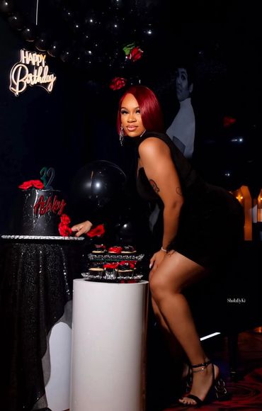 Woman in black dress posing by birthday cake and cupcakes at party.