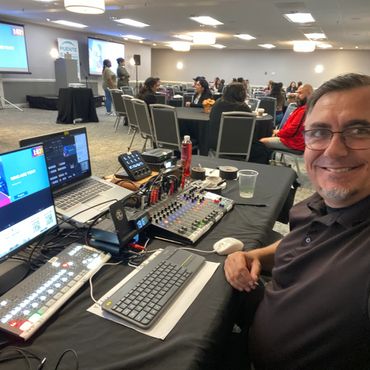 Man smiles at camera while managing audio and video equipment at an indoor event.