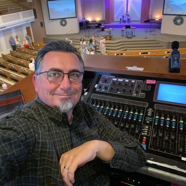 Man taking a selfie at a church sound mixing booth with people in the background.
