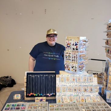 A photo of the owner Kaeden in a con-Quest Crafts t-shirt behind their table product display 