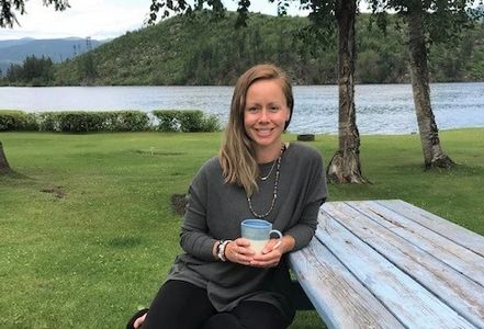 Pretty woman with sandy blonde hair, sitting at a picnic table at a park and smiling with a cup of c