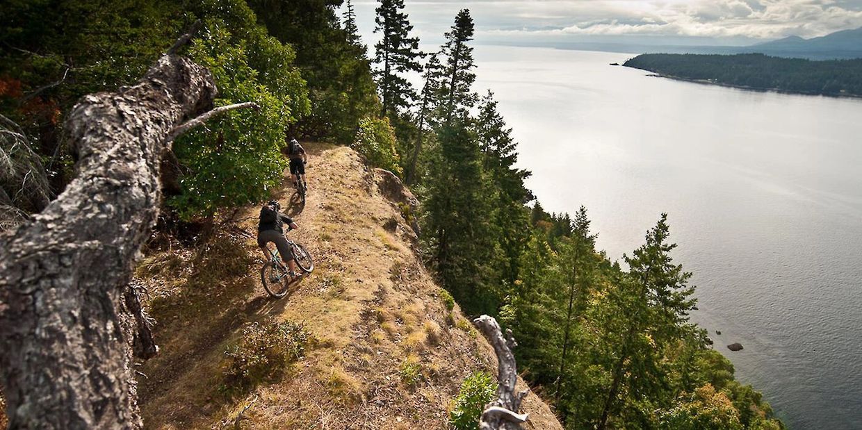 Two mountain bikers ride on a cliffside trail by the water.