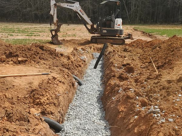 Excavator working on a trench filled with gravel and drainage pipes.