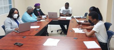A group of six people in a meeting around a conference table, some using laptops and others taking notes.