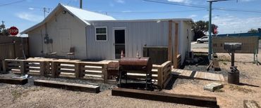 Rustic backyard with a metal grill and wooden pallets around a white building.