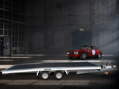 Open car trailer unladen in a warehouse with a sports car in the background.