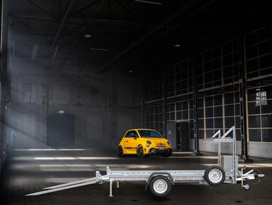 A silver lightweight car trailer with ramps down and a yellow car in the background. Single axle.