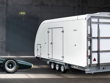 Covered car trailer in white, in a warehouse with a green sports car next to the trailer.