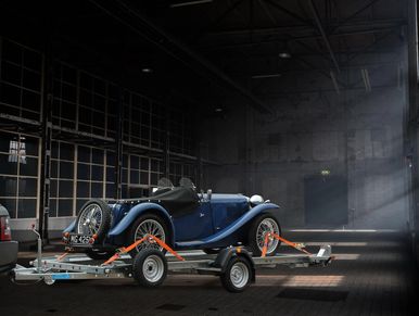 Flatbed trailer with a blue old classic car, strapped down using orange ratchet straps. Single axle.