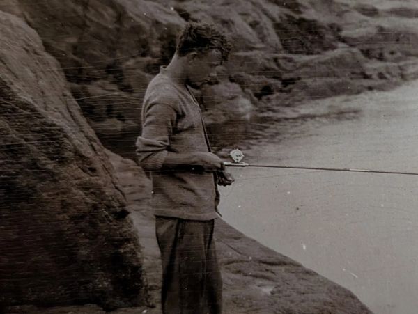 A man fishing by rocky shoreline in a vintage black-and-white photo.