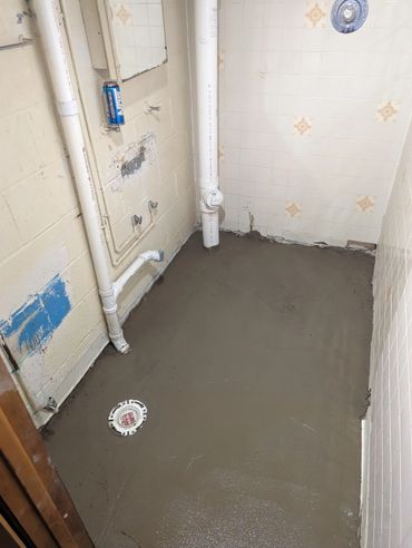 Bathroom under renovation with fresh cement floor and exposed plumbing.