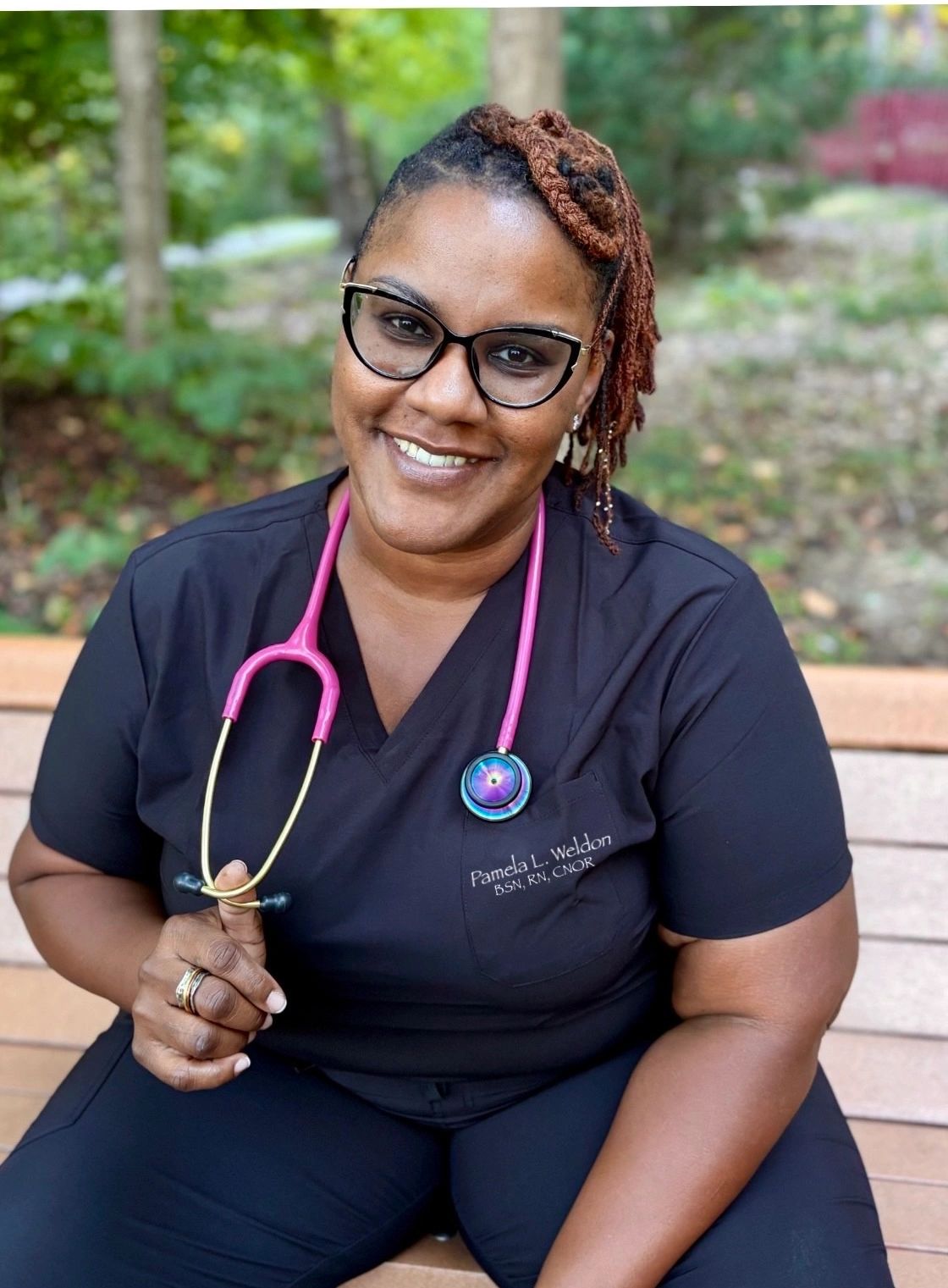 Smiling nurse with glasses and a pink stethoscope outdoors.