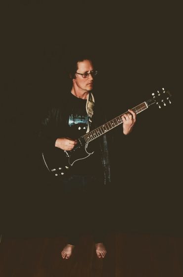 Person playing electric guitar barefoot in dim light.