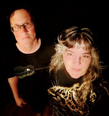 A man and woman standing against a dark background, both wearing black shirts with designs.
