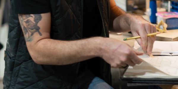 Person sketching on paper with a pencil, showing a tattooed arm and casual attire.