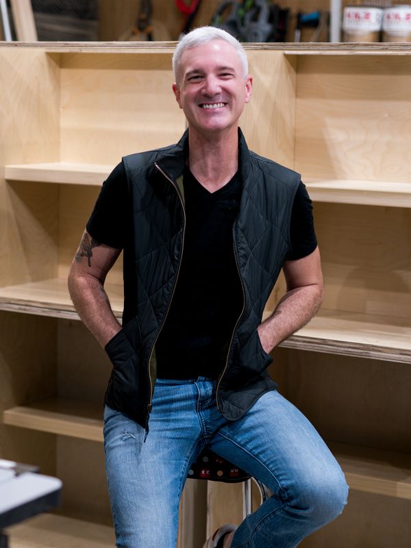 Smiling man in a black vest and jeans sitting on a stool indoors.