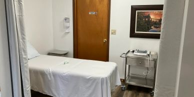 A small medical or therapy room with a bed, equipment cart, and framed picture.