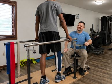 Physical therapist assists patient with leg exercises using parallel bars.