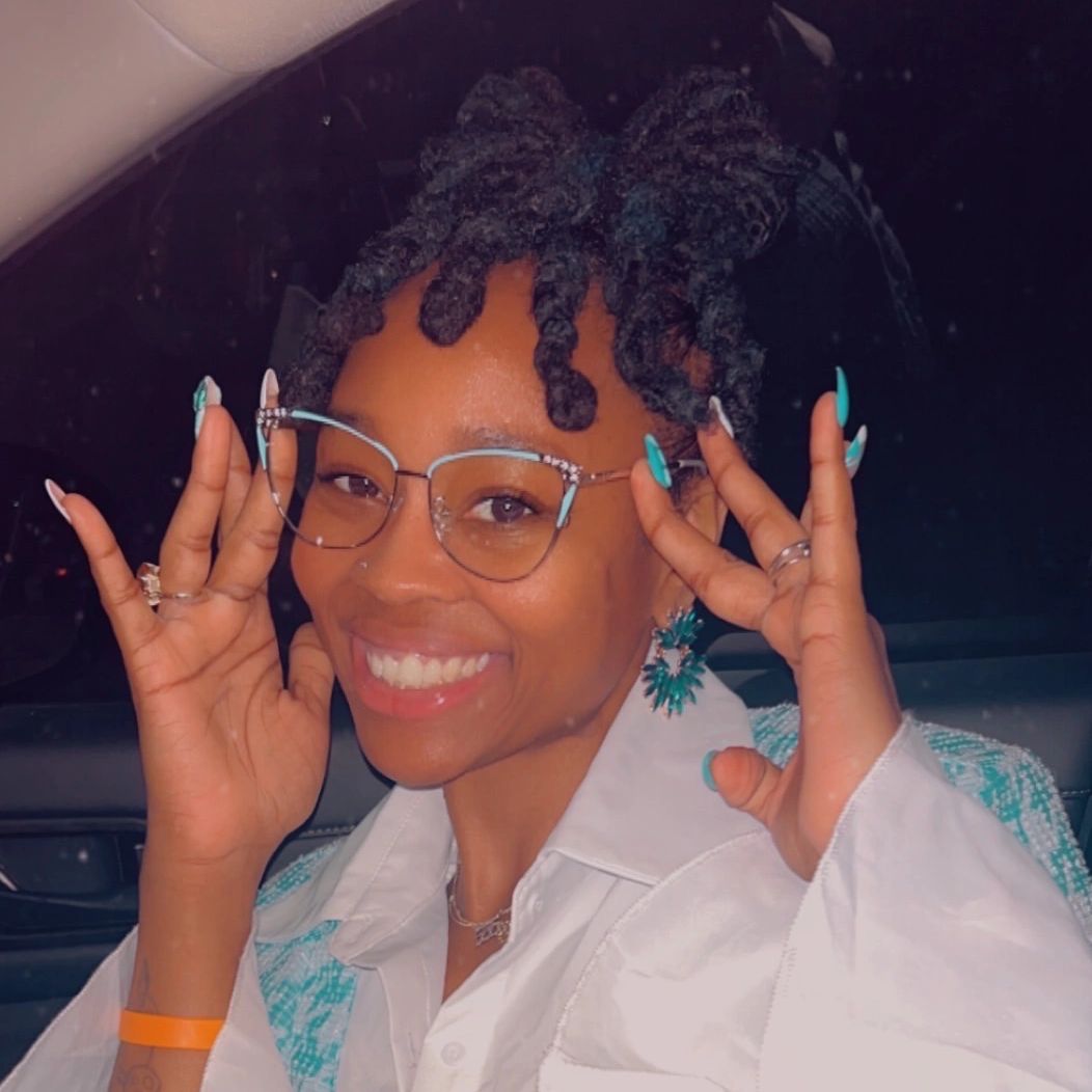 Smiling woman with glasses and stylish nails poses happily inside a car.