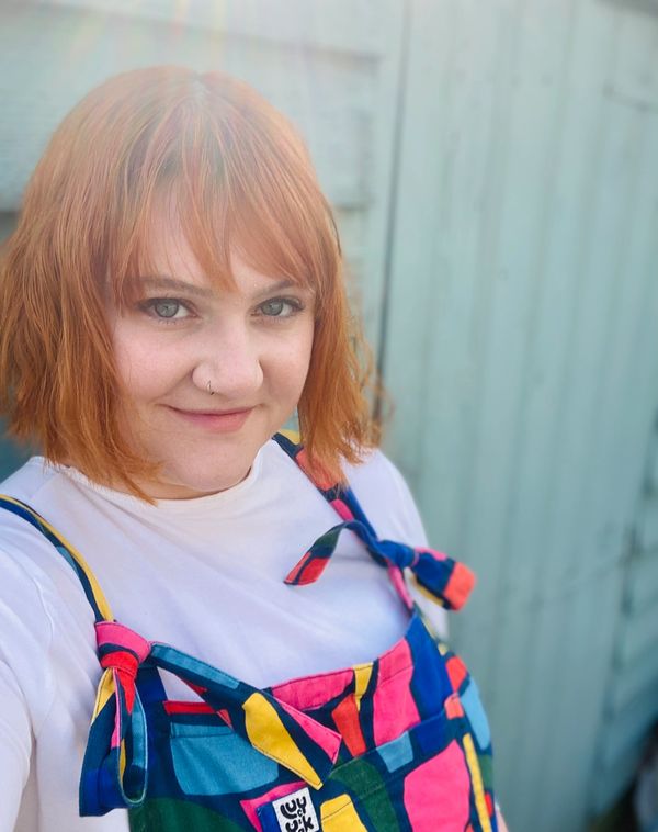 White woman with short red hair wearing bright overalls leans against a blue shed wall and smiles.