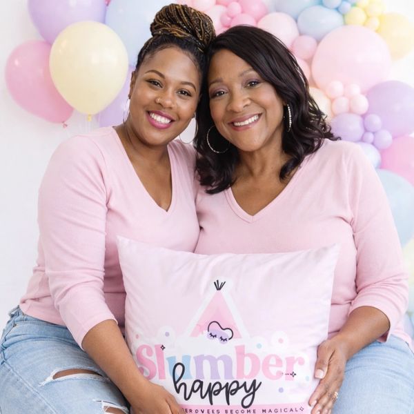 Two smiling women in pink shirts holding a pillow with balloons in the background.