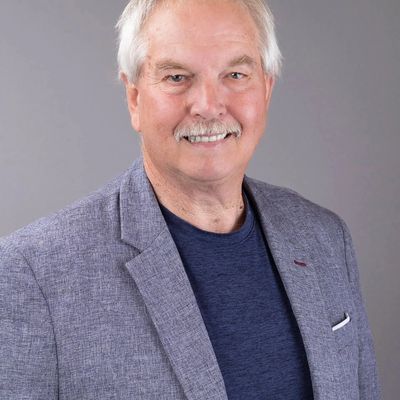 Smiling older man wearing a gray blazer and navy shirt against a gray background.