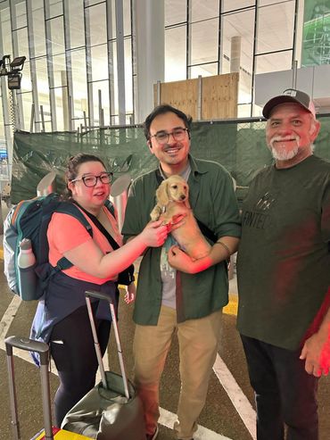 Three people smiling with a small dog at an airport or transit area.