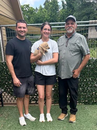 Three people smiling outdoors, woman holding a small puppy.