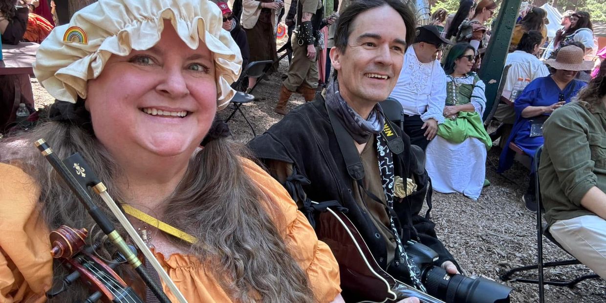 Two people smiling, one holding a violin, at an outdoor event.