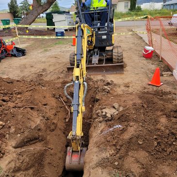 A man on a back hole digging a trench in the dirt with a orange fence around the trench