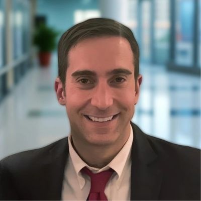 Smiling man in a suit and red tie in a bright office hallway.