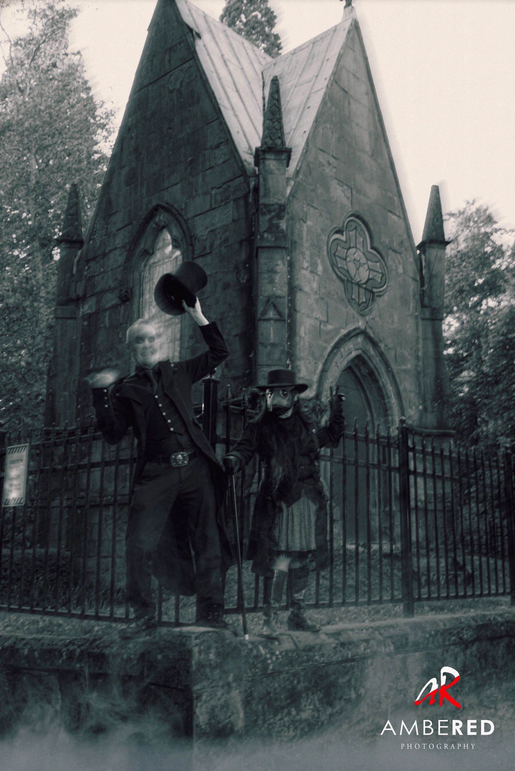 Two eerie figures in Victorian attire pose in front of a gothic stone building.