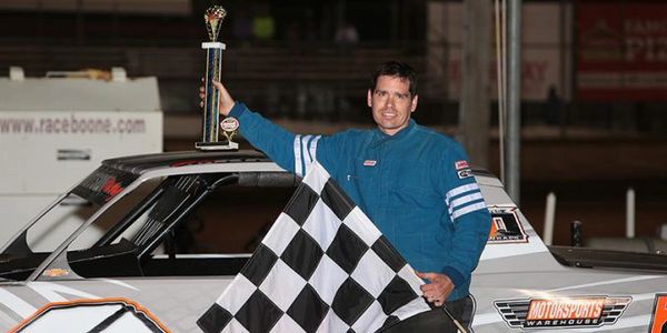 Race car driver holds trophy and checkered flag by his car.