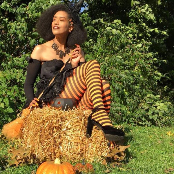 Woman in Halloween witch costume sitting on hay with pumpkins nearby.