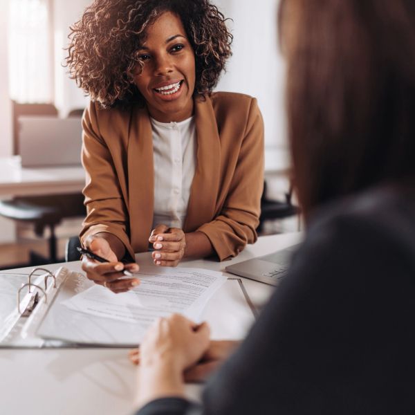 A woman in a brown blazer discusses documents with another person at a desk.