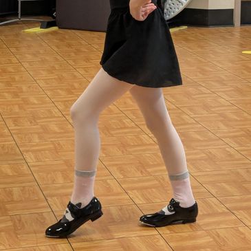Child practicing tap dance in black shoes and white tights.