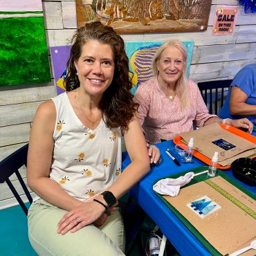 Two women smiling and sitting at a table with art supplies.