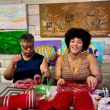 Two women crafting with red mesh ribbons at a table, colorful fish paintings behind them.