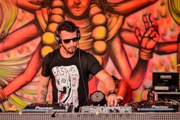 DJ wearing sunglasses and headphones performs with a colorful mural backdrop.