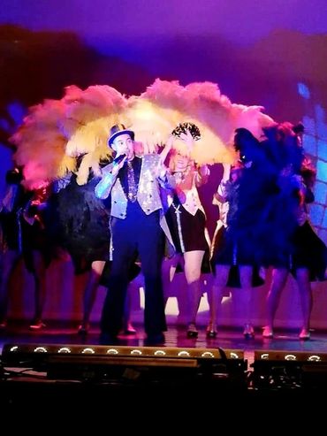 A performer sings on stage with dancers holding colorful feather fans in a lively show.