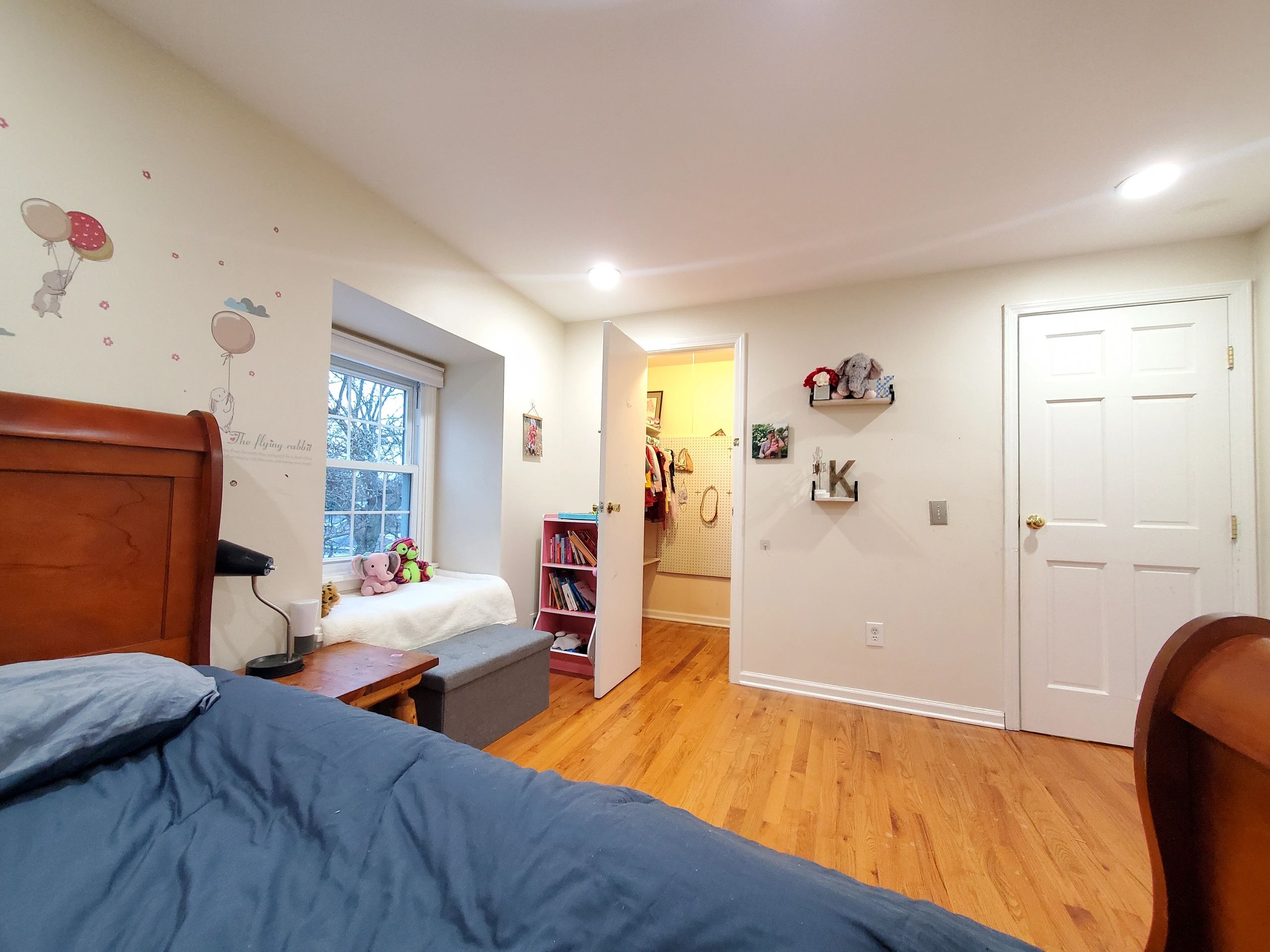 Cozy bedroom with wooden floor, stuffed animals, and a small bookshelf near a window seat.
