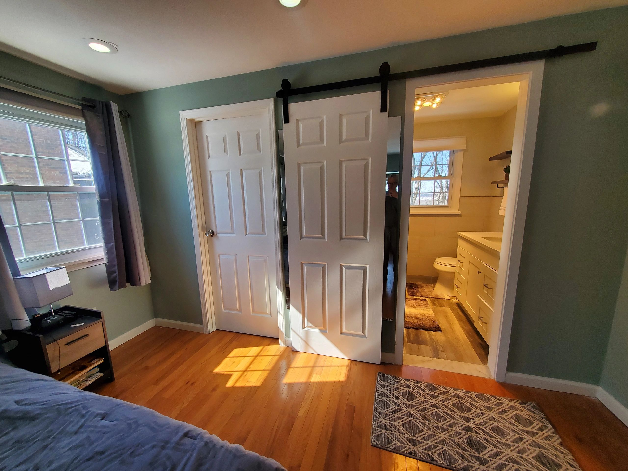 Bedroom with sliding barn door leading to bathroom, wooden floors, and window with curtains.